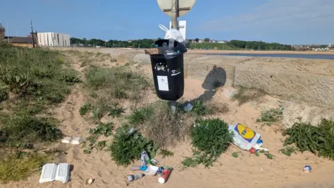 South Tyneside Council An overflowing bin attached to a signpost on the edge of a beach. Plastic bags, fish and chip boxes and bottles surround the bin.