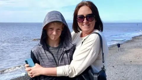 Jaime Owen A 12-year-old boy, wearing a grey zip-up jacket with the hood raised stands on a beach with shingle, sea and sand in the background. He is being embraced by his smiling mother, Jaime Owen, who is 46, has shoulder length brown hair and is wearing a white blouse and large framed sunglasses. 