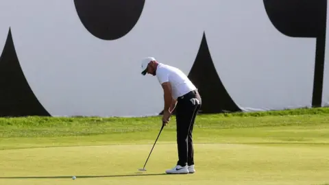 PA Media A golfer has swung back his club in an attempt to get the ball in the hole on the green. He is wearing a white baseball cap. 