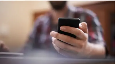 Stock image: Close-up of a person holding and looking at a smartphone, with the device in sharp focus and the background blurred. A laptop or computer is visible in the foreground, suggesting the person is using the phone while working at a desk indoors.