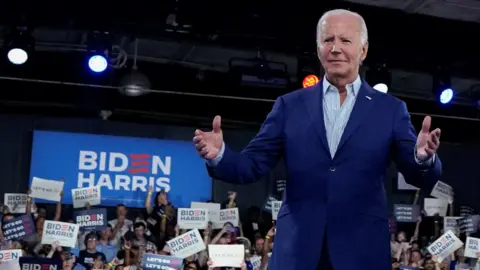 Reuters Biden mengenakan blazer biru dengan tangan terentang, di depan kerumunan dan tanda Biden Harris yang besar