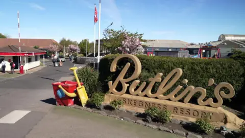 The entrance to a Butlin's resort, dominated by a large sand-coloured sign reading "Butlin's" in script and "Skegness Resort" in capital letters. Behind the sign is a tall green hedge and next to it a giant red bucket and yellow spade. In the background, a security hut, red and white raised barrier and resort buildings can be seen under a blue sky.