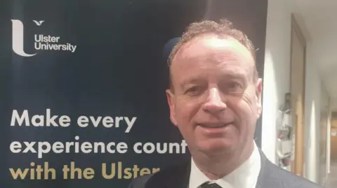 Shows a man in a dark suit and white shirt standing in front of a University Ulster placard