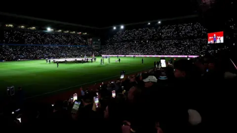 CUE Audio A football pitch at night, the pitch is floodlit and there are lots of lights in the stands coming from mobile phones being held up by fans