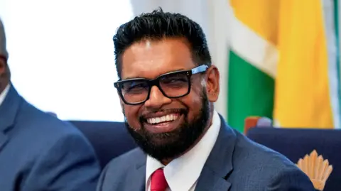 Reuters Guyanese President Irfaan Ali, wearing thick-rimmed black glasses and a navy suit, smiles widely at the camera. A Guyanese flag is draped in the background.
