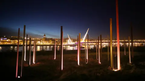 a bridge is illuminated behind a light sculpture made up of steel columns entitled on the banks of a river. a number of buildings can be seen in the background