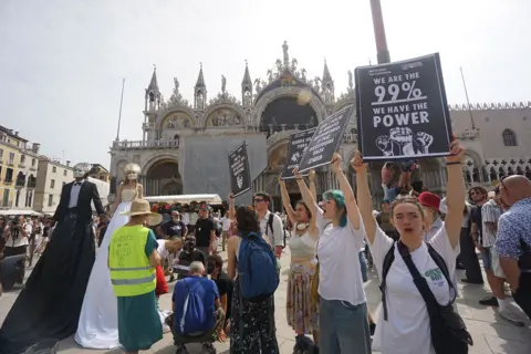Getty Images Activists protesting in Venice, Italy, against the wedding of Jeff Bezos and Lauren Sanchez. They are holding posters which read "We are the 99% We have the power". 