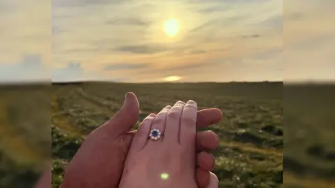 Chloe Morley Chloe Morley and Tom Heal holding hands in the field in front of the sunset. She has a round blue engagement ring on her finger