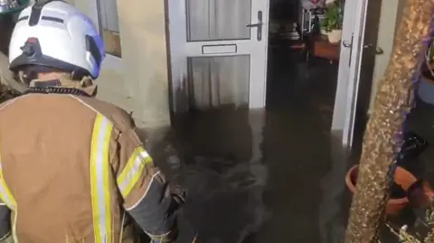 A fireman walks towards a flooded property, with water going a third of the way up the door and also in the property.