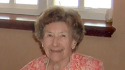 Cambridgeshire Police/PA Media Una Crown: She is smiling at the camera while sitting in front of a beige-coloured wall and window.  She is wearing a pink patterned dress with a pearl necklace and shiny earrings.