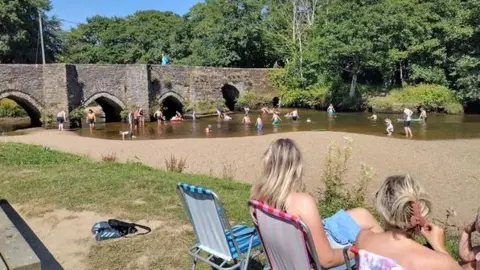 LEAF People are sitting in colourful deck chairs by the River Fowey in Lostwithiel. There are children playing in the river, there is an historic bridge with gothic arches and trees in the background. It is a sunny day and the sky is blue. The Fowey at Lostwithiel has been designated bathing water, which means the water will be tested throughout the bathing water season. 