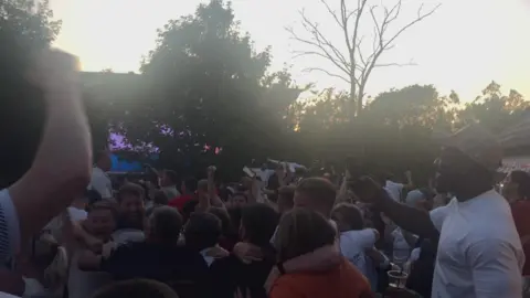 Henry Godfrey-Evans/BBC Crowds cheer, their arms outstretched, in a garden lined with trees. The lighting is dim at twilight.