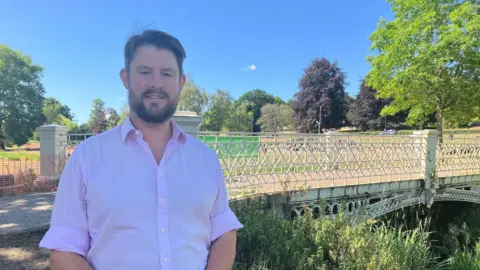 A picture of the Labour MP for Hemel Hempstead, David Taylor, who is stood in front of a white bridge in Gadebridge Park in Hemel Hempstead. He is wearing a light pink formal shirt, has short dark hair and a beard and moustache. The sky is blue behind him on a sunny day.