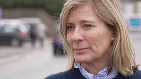 Blurred background with three cars and part of a road just about visible. Joanna Dentskevich is in the foreground on the right-hand side of the frame looking across the image to the left. She has shoulder-length blonde hair and blue eyes and is wearing a navy blue jumper. A blue and white striped frilled shirt collar is visible at the neckline of her jumper.
