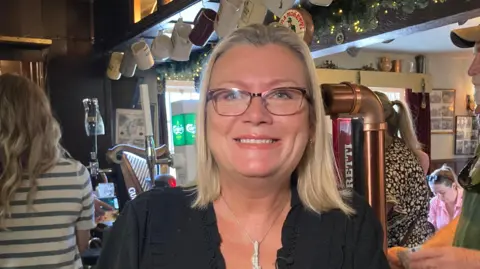 A woman wearing glasses and a black blouse stands at a pub bar counter, with shelves of liquor bottles, beer taps, hanging mugs, and garland behind them.