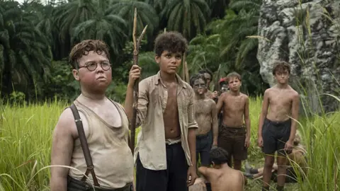 BBC/Eleven Several members of the boy cast, on an exotic island all looking very glum, ragged and dirty. Some are holding spears made from branches. Most are bare chested. Piggy in the foreground is frowning and looks puzzled