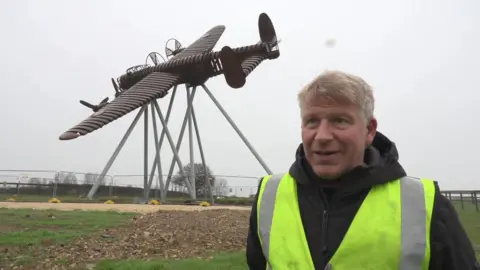 Charlie White stood in front of a life-size steel sculpture of a Lancaster Bomber. He has short blond hair and is wearing a black zip-up, hooded coat and a yellow hi-vis vest. The sky is overcast.