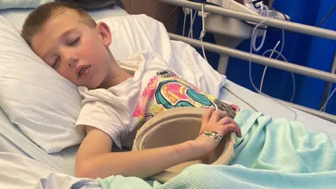 Family A boy with short hair lying asleep in a hospital bed. He is wearing a white shirt with a multi-coloured pattern on the front.