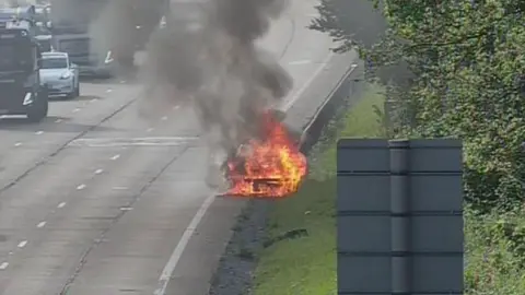 A vehicle fully engulfed in flames on the side of the road. There is a grassy bank and trees on the right.