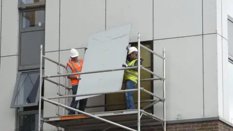 Getty Images Image showing two workers wearing white hard hats and high visibility vests removing the external cladding from Burnham Tower on the Chalcots Estate in Camden in 2017.