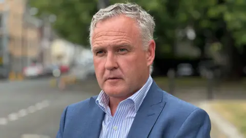 Gethin Edwards in a blue suit and light blue and white striped shirt. He has short grey hair and is standing by a road, which is blurred in the background.