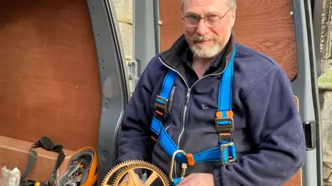 A man with glasses and a beard wearing a dark fleece and blue harness holds parts of a clock next to a van.