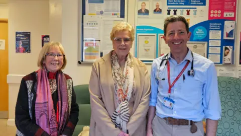 Lindel Carre, Valerie Wells and cardiology consultant Paul Williams smile at the camera. Lindel has straight shoulder-length blonde hair and wears glasses, a pink scarf and a black and white cardigan. Valerie has short blonde hair and is wearing glasses, a beige coat and a white scarf with orange flowers.  Paul Williams has short brown hair and is wearing a blue shirt and beige trousers. There is a stethoscope over his neck.