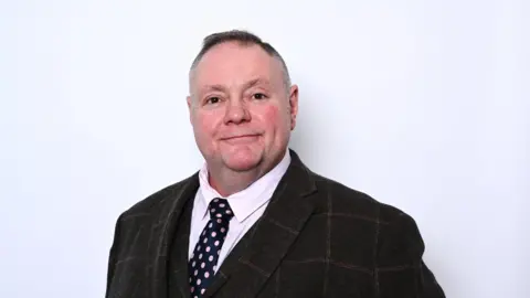 Labour Council leader Stephen Simkins, wearing a brown check suit and tie standing against a white background