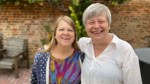 BBC Two female friends smiling with their arms around each other outside in a garden. There is a brick wall and a garden chair behind them.