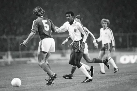 Shutterstock A black and white image of Viv Anderson, wearing a white long-sleeved shirt, dark shorts and white socks, during his England debut. A Czechoslovakia player wears a dark shirt bearing the number 5. His shorts are white.