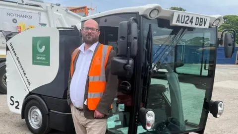 Paul Wells Councillor Paul Wells from Great Yarmouth Borough Council stood in front of a road sweeper vehicle. He is bald and has a beard and black wide-framed glasses. He is wearing a hi-vis jacket over the top of his suit.