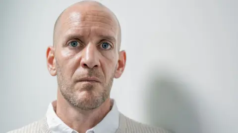 Dan Barr, 41, who has blue eyes, a bald head and light stubble, speaks into the camera wearing a beige jumper over a white shirt.