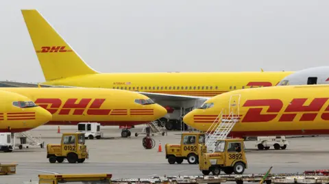 Reuters Four yellow planes with DHL's red lettering on them on a runway. Three planes are facing right, one is facing left. There are three small, yellow single-seater airport vehicles on the runway.