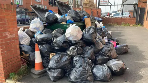 Black bin bags overflowing from a large green bin onto the pavement. They are piled up both on top of an around the bin. 