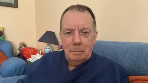 A man with short black hair wearing a blue top. He is sat in a living room with blue sofas. The walls behind him are cream. He is looking into the camera
