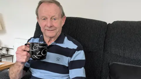 A man in his eighties sits on a black sofa in a living room. He is holding a black coffee mug. A lampshade is in the background.