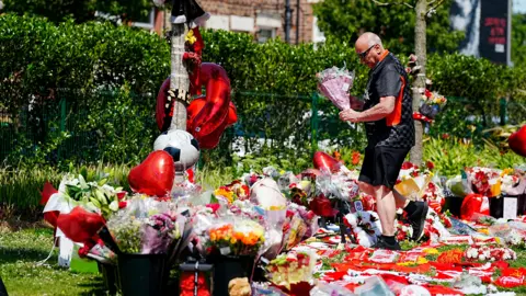 PA Media A man in a black t-shirt, black shorts and sunglasses stands amid a sea of tributes including flowers, football shirts, balloons and flowers with a bouquet in his hand