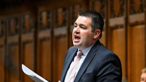 Reuters MP Shaun Davies, holding paper and wearing a blue suit, addressing the House of Commons, with wooden panelling behind him