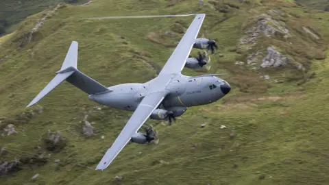 RAF The A400M in the air, flying over a green, rocky, hilly region.