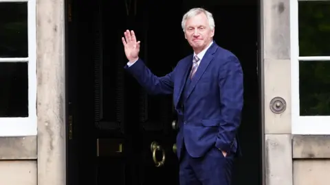 PA Media Ivan McKee, wearing a blue checked suit and a pink striped tie waves as he stands in front of a black door.