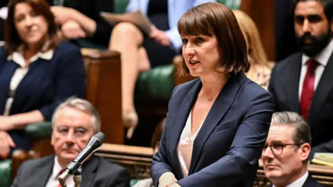 Chancellor Rachel Reeves is pictured speaking at the dispatch box in the House of Commons. She wears a blue suit jacket, white blouse and delicate necklace. 