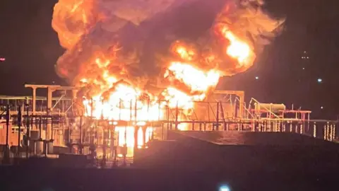 PA Media Black smoke and large yellow flames billow into the night sky in between power cables at the electrical substation. Rows of metal fences can be seen in the foreground