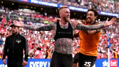 PA Media A footballer, with his top off exposing a sports vest, holds both hands up in celebration as a team-mate reaches out to him. Behind them fans in the terraces go wild.