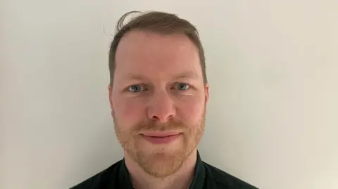 Javed Love is looking directly at the camera. He has a ginger beard and short light brown hair. 