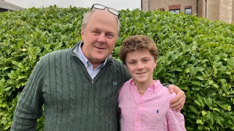Tom is wearing a pink shirt with a blue ralph lauren logo.He has curly hair and is standing in front of a bush, looking at the camera smiling. He is being hugged from the side by his dad, Greg, who is wearing a green knit jumper. He has his glasses tucked behind his ears, balancing on the top of his head.