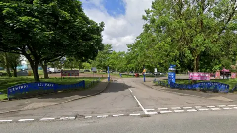 The entrance to a car park, which has blue signage indicating it is Central Forest Park. There are white dashed give way markings at the junction with the road, and the car park is surrounded by trees.
