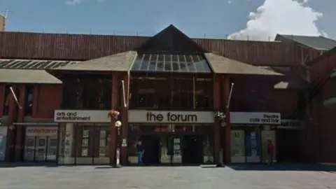 Google A street view of The Forum which is a large brown brick building with a pointed roof above the entrance. The sign above the door reads 'the forum' in lowercase letters with the words 'arts and entertainment' and 'theatre cafe and bar' on either side.