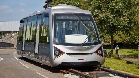 Coventry City Council A modern silver train carriage on a length of track. There is an area of open space next to the track with grass and trees and a man is standing there.