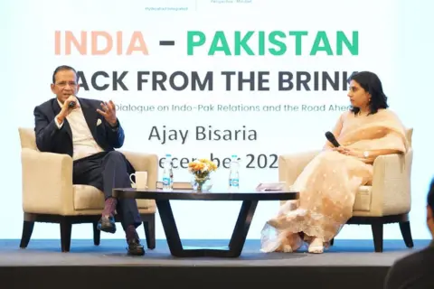 Ajay Bisaria Ajay Bisaria duduk di podium memegang mic di tangannya.