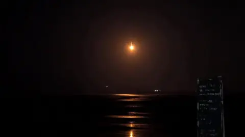 Image shows a near pitch black beach with the distant light in the sky, potentially from a police helicopter or a flare.
